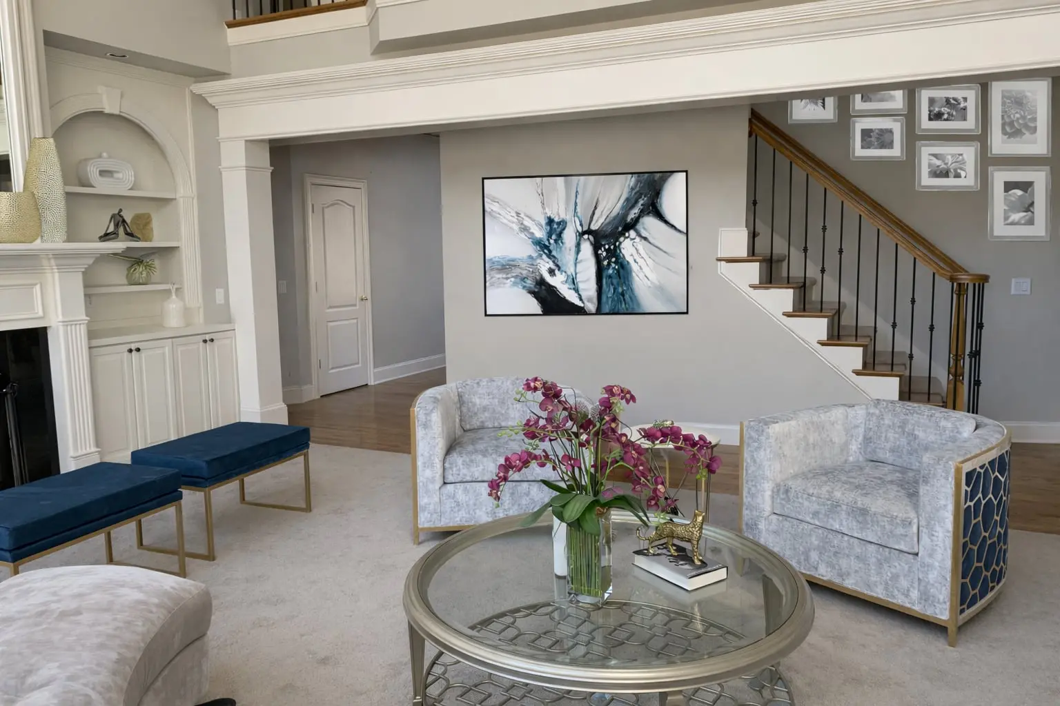 Grand two story living room with abstract art above seating area, blue velvet benches, glass coffee table, and staircase gallery wall designed by Welcome Home Interiors in Hoschton Georgia