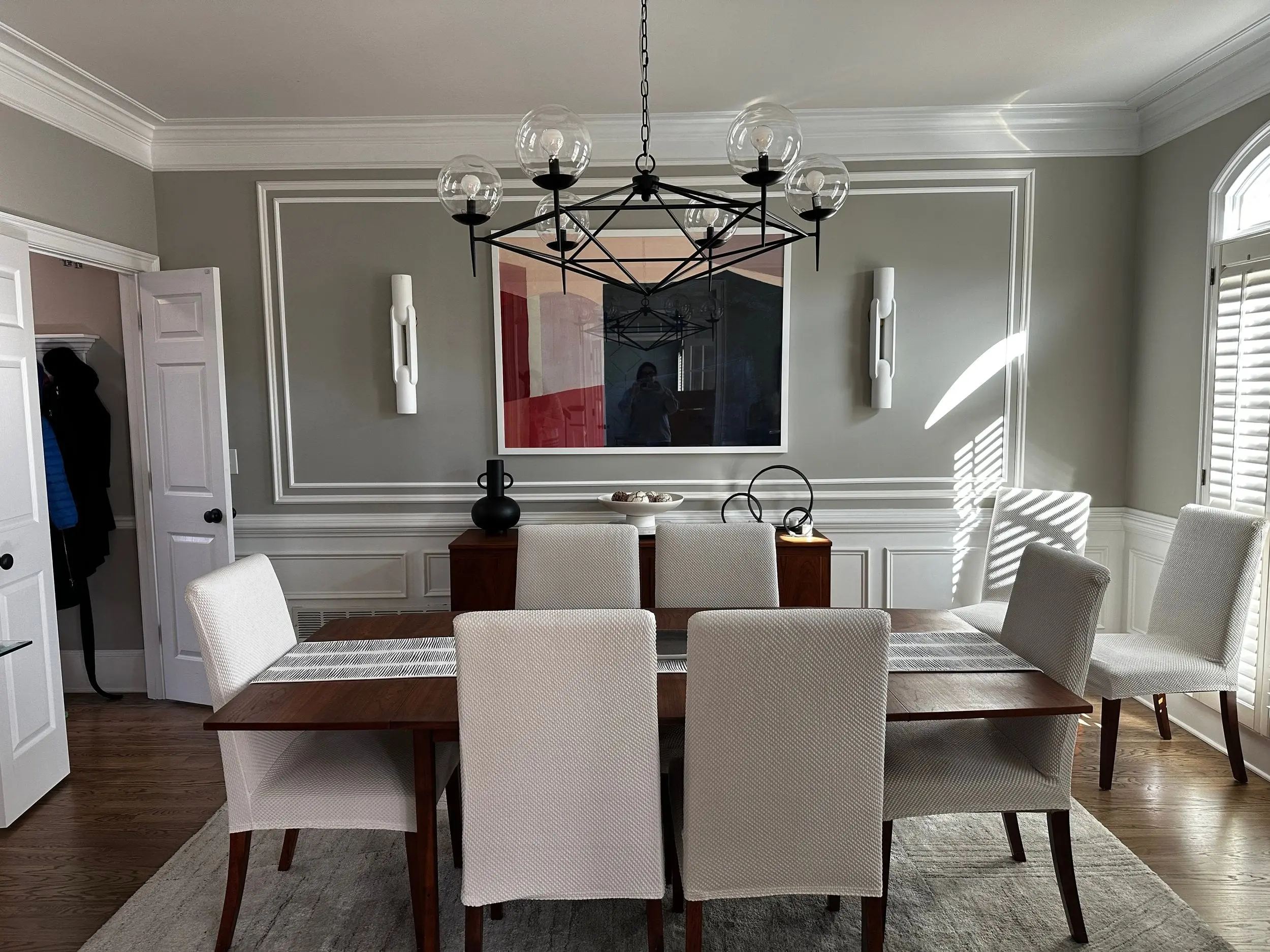 Dining room with gray walls, white wainscoting, a black geometric chandelier and light upholstered chairs